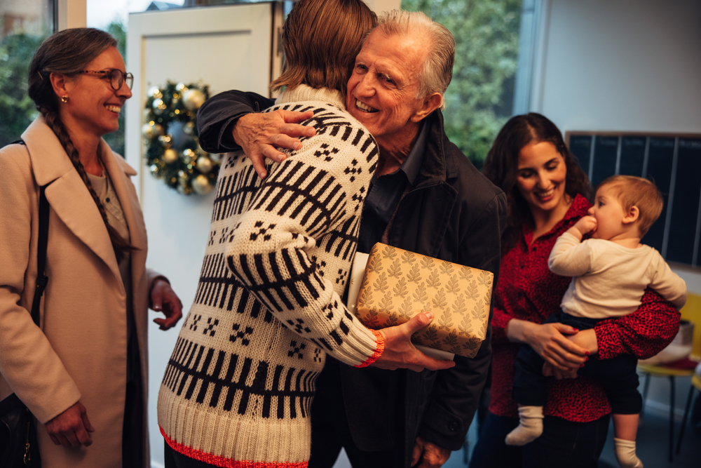 Guests arriving at home for holidays. There's a man hugging an elderly person while holding Christmas gifts in the center, with a mother and her baby on the right and another happy woman on the left.